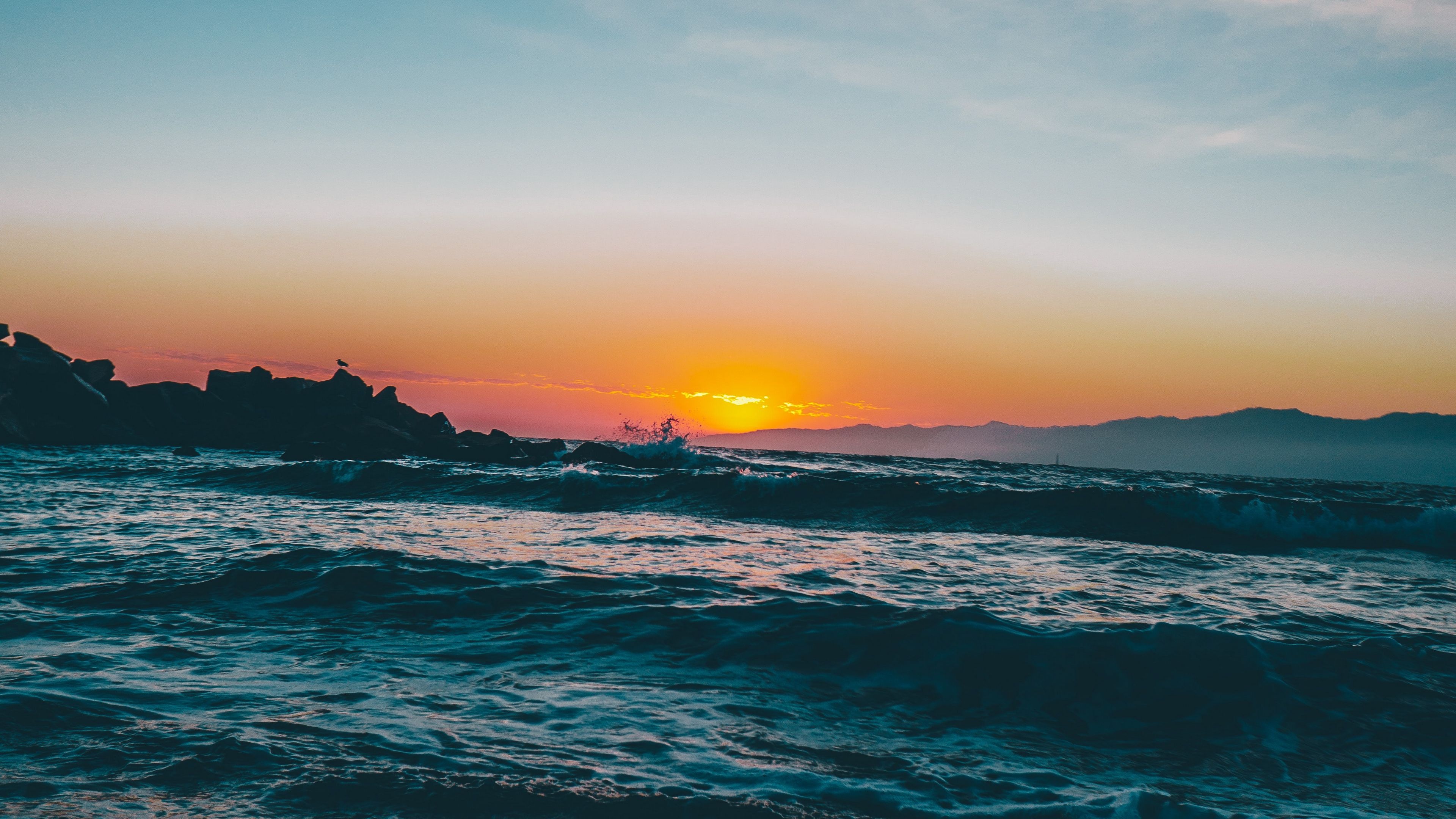crashing ocean surf waves, rocky jetty silhouette, sunset over coastal mountains, 4K Desktop wallpaper