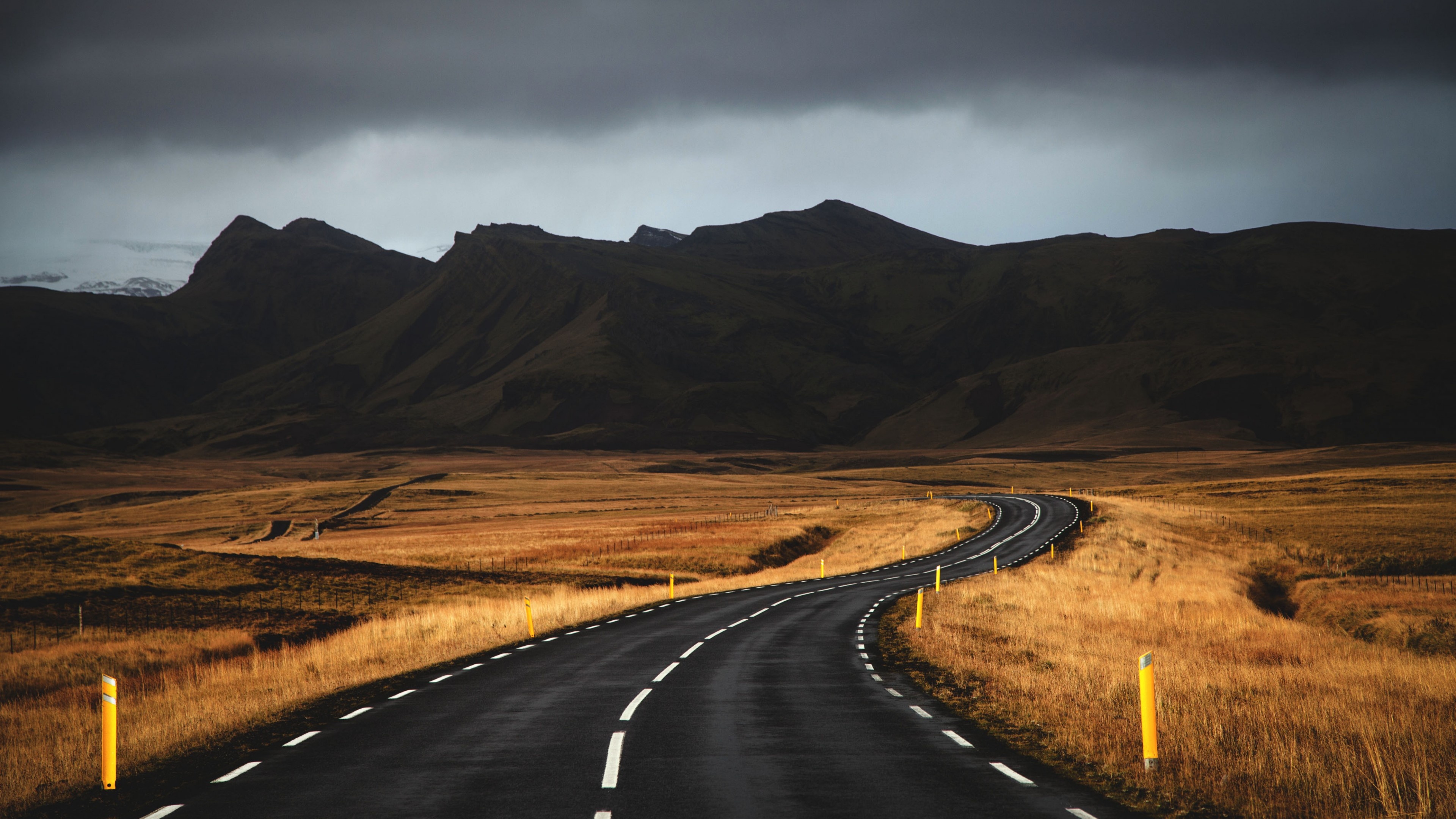 curving asphalt road, yellow roadside marker posts, dark volcanic mountains, 4K Desktop wallpaper